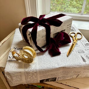 May include: A rectangular gift box wrapped in white paper with black printed newspaper-style text, tied with a deep burgundy velvet ribbon. A gold-toned tape dispenser and a pair of gold-toned scissors are placed on top of the wrapped gift.