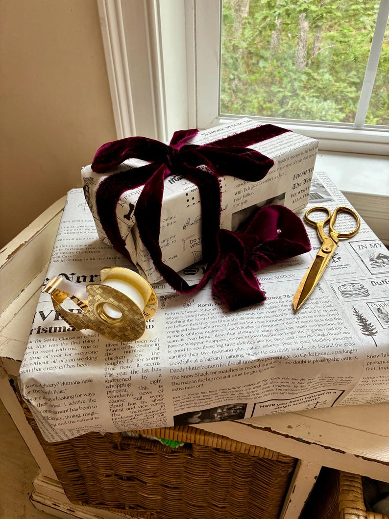 May include: A rectangular gift box wrapped in white paper with black printed newspaper-style text, tied with a deep burgundy velvet ribbon. A gold-toned tape dispenser and a pair of gold-toned scissors are placed on top of the wrapped gift.