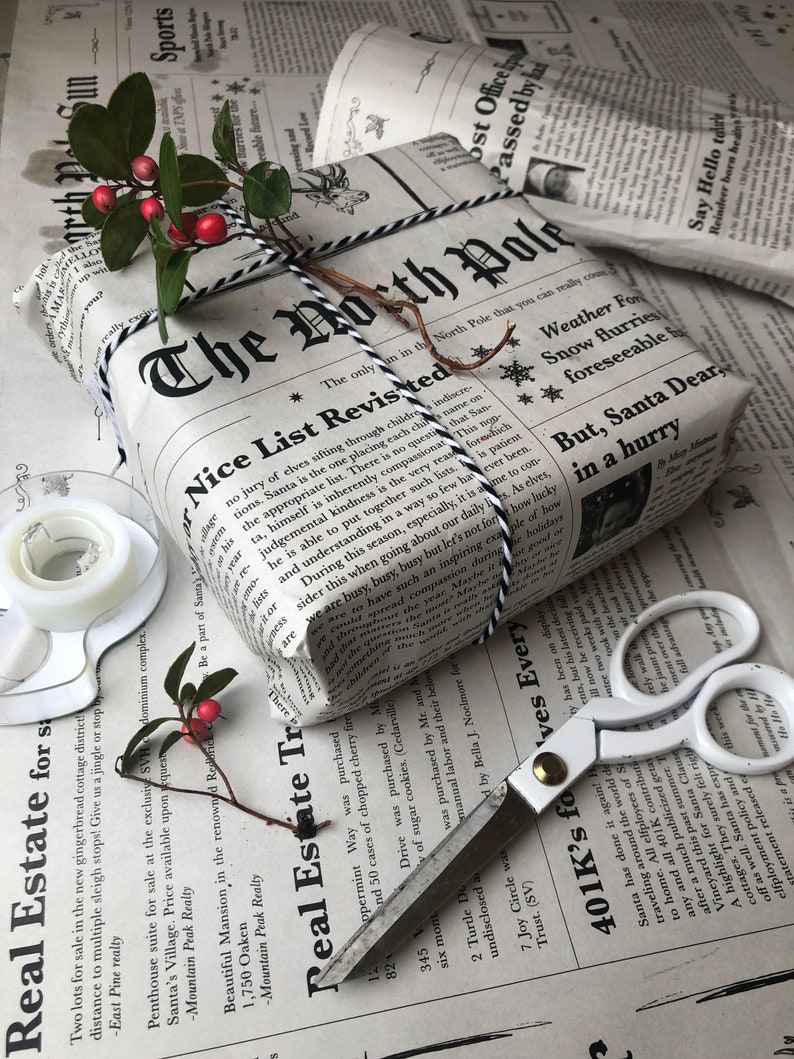 May include: A rectangular gift wrapped in newspaper-style paper with the headline The North Pole Nice List Revisited. The package is tied with black and white baker's twine and topped with a sprig of green leaves and small red berries.