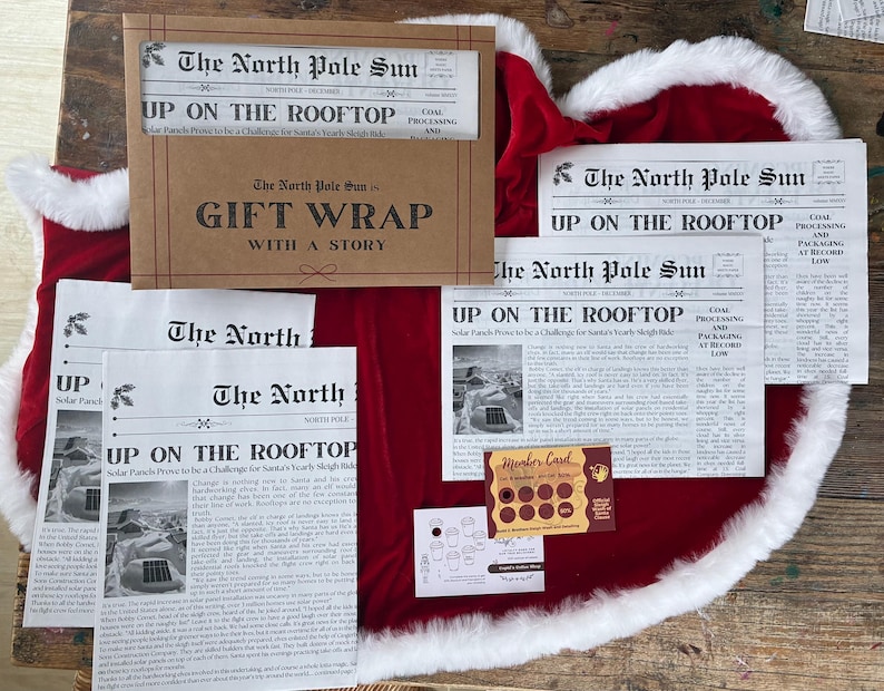 May include: A set of gift wrap sheets featuring The North Pole Sun newspaper print with the headline Up On The Rooftop, a brown envelope, a small white card with coffee cup icons, and a brown scratch-off member card with circular gold foil spots.