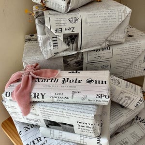 May include: A stack of rectangular gift boxes wrapped in white paper printed with black newspaper-style text and illustrations. The top box is adorned with a gold star garland, while a lower box features a soft, dusty pink velvet ribbon tied in a bow.