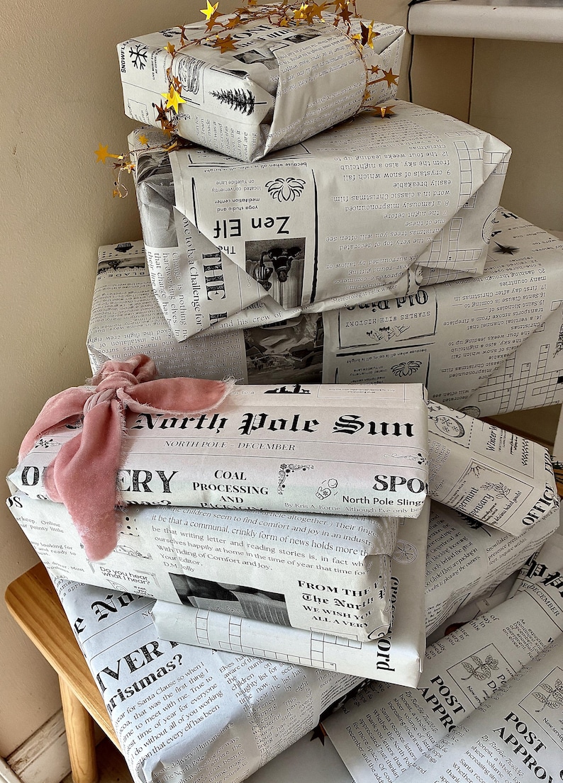 May include: A stack of rectangular gift boxes wrapped in white paper printed with black newspaper-style text and illustrations. The top box is adorned with a gold star garland, while a lower box features a soft, dusty pink velvet ribbon tied in a bow.