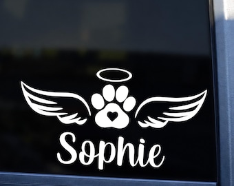 May include: A white vinyl decal applied to a vehicle window featuring a central paw print with a small heart in the center, flanked by two stylized wings and topped with a halo. The name Sophie is printed in a white cursive script below the graphic.