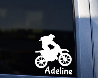 May include: A white vinyl decal applied to a vehicle window, featuring a silhouette of a person riding a motocross dirt bike. Below the bike, the name Adeline is written in a white, stylized, cursive-inspired font.