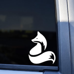 May include: A white vinyl decal featuring a stylized, minimalist silhouette of a sitting fox with a curved, bushy tail. The graphic design is applied to the dark tinted glass of a vehicle window, creating a high-contrast visual effect.