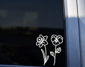 May include: A white vinyl decal featuring a line art illustration of a daffodil and a pansy flower. The decal is applied to the dark tinted rear window of a white vehicle, displaying the two distinct floral outlines in a clean, simple style.