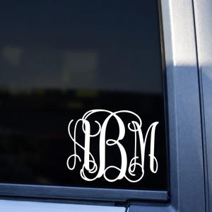 May include: A white vinyl decal featuring a decorative, interlocking monogram with the letters A, B, and M in a stylized script font. The decal is applied to the dark glass of a vehicle window, displaying a clean, high-contrast design.