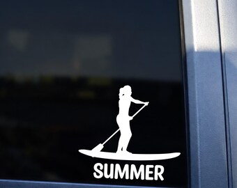 May include: A white vinyl decal applied to a vehicle window featuring a silhouette of a person standing on a paddleboard while using a paddle, with the word SUMMER printed in bold white capital letters directly underneath the graphic.
