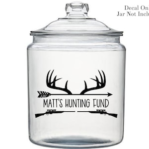 May include: A clear glass jar with a black and white decal that says "Matt's Hunting Fund". The decal features a pair of antlers, an arrow, and two rifles.