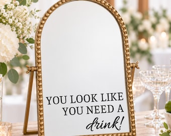 Op de afbeelding: Een boogvormige tafelspiegel met een goudkleurige kralenrand. Het glasoppervlak is voorzien van zwarte tekst die luidt: "YOU LOOK LIKE YOU NEED A drink!". De spiegel staat op een goudkleurige metalen standaard met verstelbare knoppen aan de zijkant.