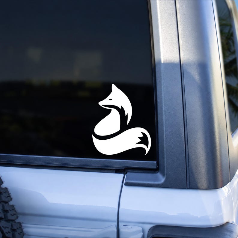 May include: A white vinyl decal featuring a stylized, minimalist silhouette of a sitting fox with a curved, bushy tail. The graphic design is applied to the dark tinted glass of a vehicle window, creating a high-contrast visual effect.