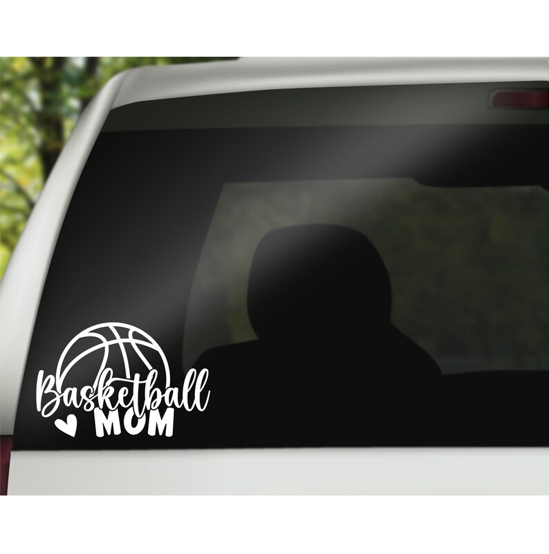 May include: A white vinyl decal applied to a car window featuring a line-art basketball graphic above the script text Basketball and the block-letter text MOM, with a small heart icon positioned to the left of the word MOM.