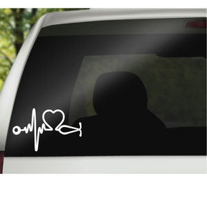 K&ouml;nnte beinhalten: Wei&szlig;er Vinyl-Aufkleber mit einem Herzen, einem Stethoskop und einer Herzschlaglinie. Der Aufkleber befindet sich auf der Heckscheibe eines Autos.