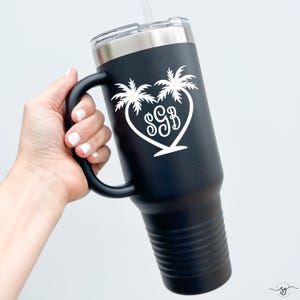 May include: Black insulated tumbler with a handle, a silver lid, and a white straw. The tumbler features a white heart-shaped design with palm trees and the initials "SGB". The tumbler is held by a hand with white nail polish.
