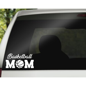 May include: A white vinyl decal applied to a vehicle window featuring the text Basketball MOM in a bold, block-style font. The letter O in the word MOM is replaced by a graphic illustration of a basketball with visible black seam lines.