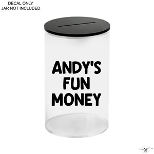 Op de afbeelding: Een doorzichtige cilindervormige houder met een zwarte deksel voorzien van een horizontale gleuf. De voorzijde toont de zwarte tekst ANDY'S FUN MONEY. Links bovenaan staat DECAL ONLY JAR NOT INCLUDED.