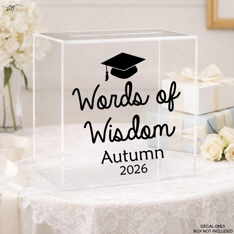 May include: Clear acrylic box with a slot on top, featuring black text that reads "Words of Wisdom Autumn 2026" and a graduation cap graphic. The box is on a white lace tablecloth, with flowers and wrapped gifts in the background.