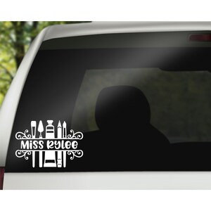 May include: White vinyl decal with the text "Miss Kylee" and a design of paintbrushes, paint tubes, and a palette knife. The decal is on a black car window.