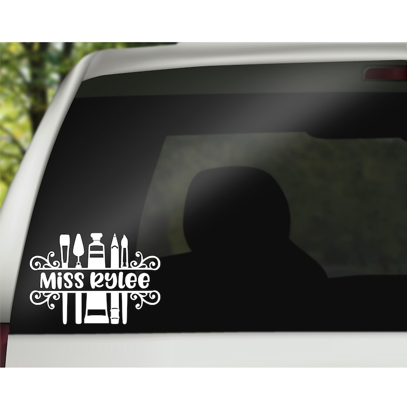 May include: White vinyl decal with the text "Miss Kylee" and a design of paintbrushes, paint tubes, and a palette knife. The decal is on a black car window.