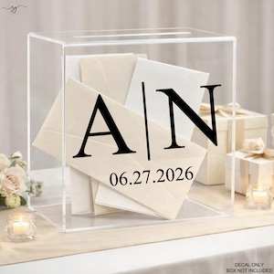 May include: Clear acrylic wedding card box with black lettering featuring the initials "A | N" and the date "06.27.2026". The box is filled with cream-colored envelopes.