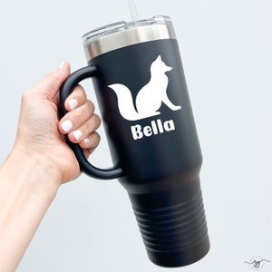 May include: A matte black stainless steel tumbler with a handle, a clear lid, and a straw. The tumbler features a white silhouette graphic of a sitting fox above the name Bella printed in a bold, white, sans-serif font on the side.
