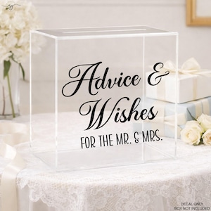 May include: Clear acrylic box with a slot on top, featuring black script that reads "Advice & Wishes FOR THE MR. & MRS." Ideal for weddings or events, the box sits on a white lace tablecloth, with flowers and gifts in the background.