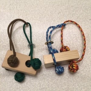 Puede incluir: Dos rompecabezas de madera. Uno tiene una base de madera marrón claro con cuerdas anudadas verdes y marrones. El otro tiene una base de madera marrón claro con cuerdas anudadas azules, naranjas y amarillas. Ambos tienen anillos metálicos.