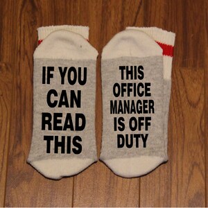 May include: A pair of grey and white socks with black text. One sock reads "If you can read this" and the other sock reads "This office manager is off duty".