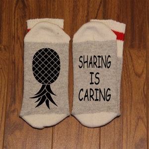 May include: A pair of white socks with grey stripes. One sock has a black pineapple design and the other sock has the text "Sharing is Caring" in black.