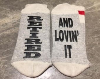 May include: A pair of gray ankle socks with the words "Retired" and "And Lovin' It" printed on them. The socks have a red stripe on the heel.