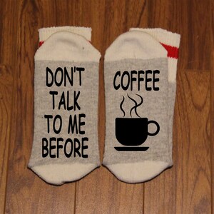 Puede incluir: Un par de calcetines grises y blancos con rayas rojas. Un calcetín dice "No me hables antes del café" y el otro calcetín tiene una taza de café humeante.