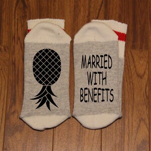 May include: A pair of grey and white socks with red stripes. One sock has a pineapple graphic and the other sock says "Married with Benefits".
