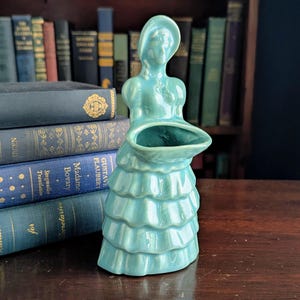 May include: A turquoise ceramic figurine of a woman wearing a hat and a tiered dress. The figurine has a large opening in the front, likely for holding flowers or other small items. The piece is displayed on a wooden surface with a backdrop of books.