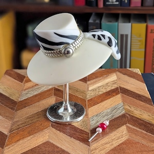 May include: A miniature white hat with a black zebra print band and a silver-toned metallic ring accent, mounted on a silver-toned pedestal stand. Beside the stand lies a small silver-toned pin featuring two white beads and one central red bead.