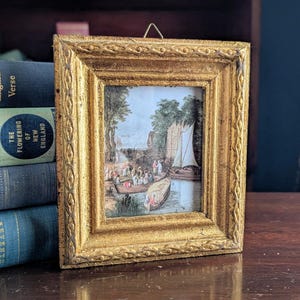 May include: A small, antique framed print with a gold-colored, ornate frame. The print depicts a river scene with boats, people, and trees. The frame has a decorative, textured surface. The print is displayed on a wooden surface next to a stack of books.