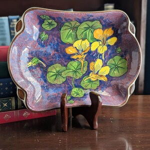May include: A decorative, square-shaped ceramic dish with a scalloped edge. The dish features a floral design of yellow flowers and green leaves against a mottled blue and purple background. The dish is displayed on a wooden stand.