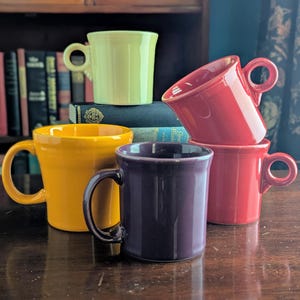 May include: A collection of five ceramic mugs in various solid colors including light yellow, bright red, deep yellow, and dark purple. Each mug features a smooth, glossy finish, a cylindrical body, and a rounded handle attached to the side.