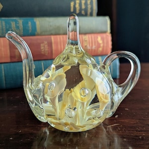 May include: A clear glass teapot figurine featuring an internal design of pale yellow calla lily flowers suspended within the glass. The teapot has a curved spout, a rounded body, a pointed lid finial, and a handle, all crafted from transparent glass.