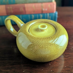May include: A round, yellow ceramic teapot featuring a distinct crackle glaze finish. The teapot has a single curved handle on the side and a matching circular lid with a small, centered knob handle on top, showcasing a glossy, textured surface.