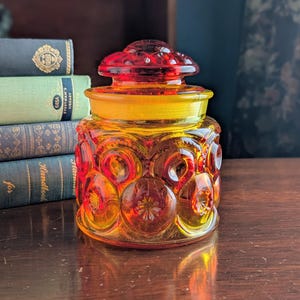 Op de afbeelding: Een vintage amberkleurige glazen pot met een deksel. De pot heeft een sterrenpatroon en een ronde vorm. De deksel is donkerder amberkleurig. De hoogte is ongeveer 15 cm.