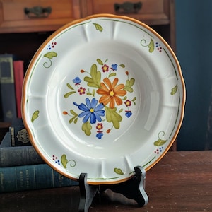 May include: White ceramic serving bowl with a floral design. The bowl features a scalloped edge with an orange trim. The center of the bowl has a bouquet of orange, blue, and red flowers with green leaves. The bowl is displayed on a black stand.