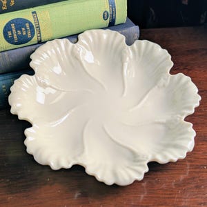 May include: Cream-coloured, leaf-shaped ceramic dish. The dish features a scalloped edge and a central design of radiating veins. The dish is on a wooden surface, with books in the background.