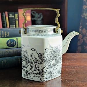 Puede incluir: Una tetera de porcelana blanca con una escena pastoral en blanco y negro. La tetera tiene un asa y un pico dorados. El borde superior está decorado con un motivo floral negro. La tetera es rectangular y se apoya sobre una superficie de madera.