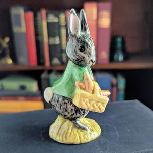 May include: A ceramic figurine of a rabbit with grey fur, wearing a green jacket, and carrying a small yellow woven basket filled with orange carrots. The rabbit stands on a textured yellow base, depicted in a side profile view with upright ears.