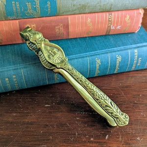 May include: A gold-toned metal nutcracker featuring an embossed castle design on the central hinge and intricate leaf-like scrollwork patterns along the length of the handles. The tool has a polished finish and a hinged mechanism for cracking nuts.