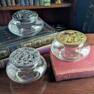 May include: Three clear glass jars with decorative lids. One jar has a silver-colored lid with a floral design, another has a gold-colored lid with a floral design, and the third has a silver-colored lid with a leaf design. The jars are on top of antique books.