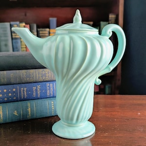 May include: A light blue ceramic teapot with a unique, swirling design. The teapot has a lid and a curved handle. The base is a pedestal shape. The teapot is displayed on a wooden surface with antique books in the background.