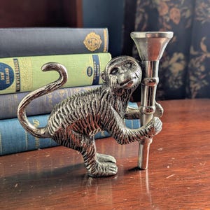 May include: A silver-colored monkey figurine holding a metal candlestick. The monkey has detailed fur texture and is posed with its tail curled. The candlestick has a flared top and a cylindrical stem. Books are in the background.