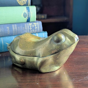 May include: A brass frog-shaped trinket box with a hinged lid. The box is a warm, antique gold color and sits on a dark wooden surface. A stack of vintage books in blue and green are in the background.