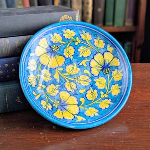 以下が含まれることがあります： 鮮やかな花柄が描かれた、手描きの丸いセラミックプレート。明るいターコイズブルーの背景に、黄色の花と緑の葉が描かれています。縁は濃いターコイズブルーです。装飾的な食器。